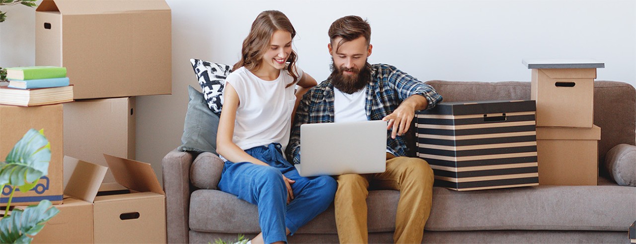 Young couple sitting on couch in process of moving. They are looking at mortgage rates on a laptop.