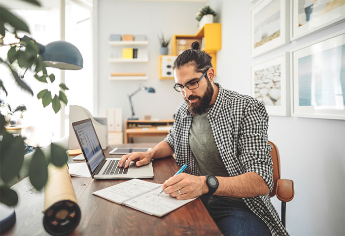 Bearded man with glasses at desk using laptop and writing