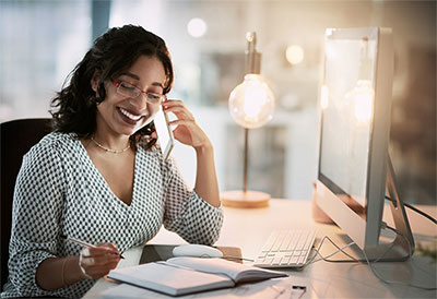 Woman at computer desk writing and talking on cellphone.